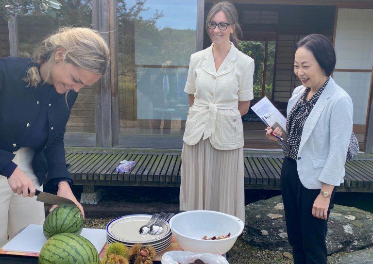 Photo of a woman slicing watermelon with 2 other women watching.